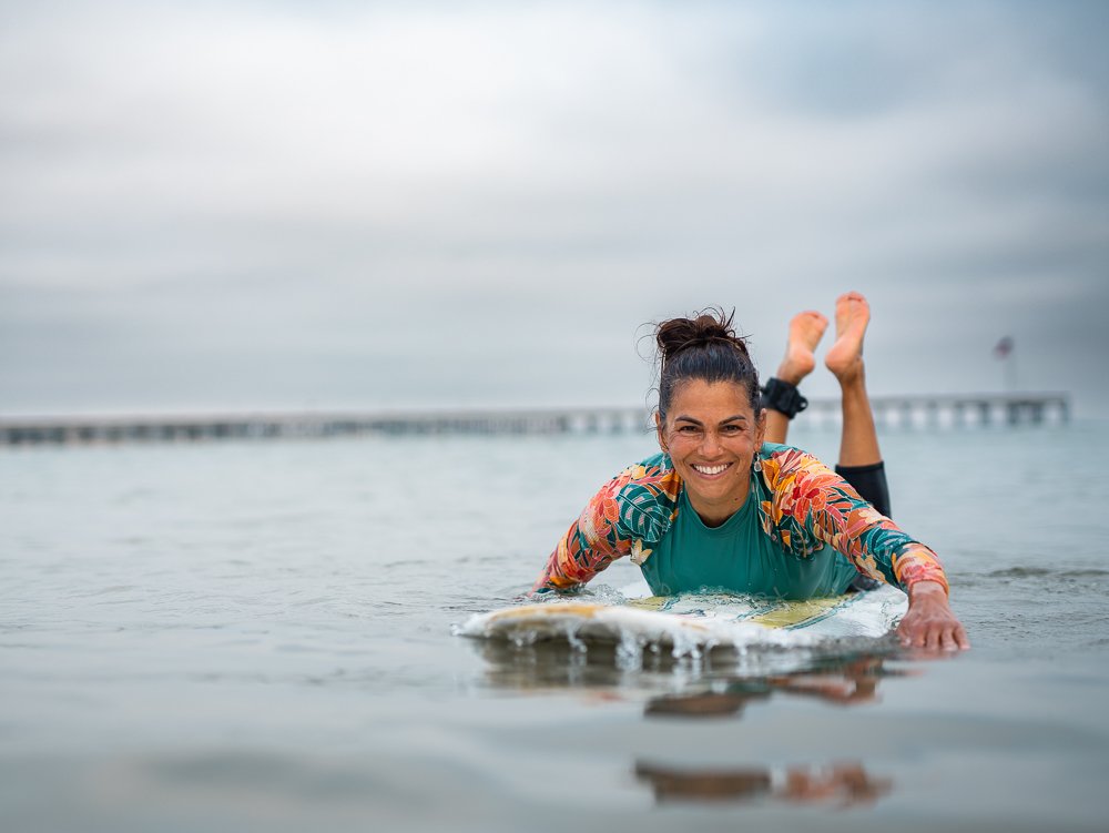 Dr. Kavitha Simpson smiling from her surfboard in the ocean near a pier