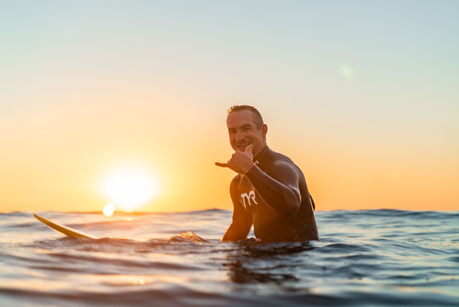Dr. Jamey Sotis sitting on his surfboard at sunset, waving to the camera